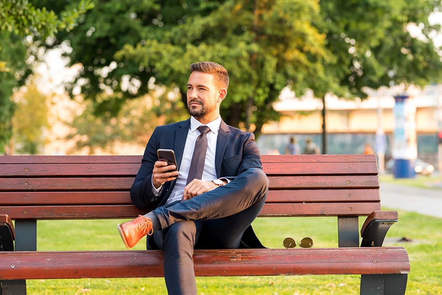Client Center - Businessman Sits on a Park Bench With Coffee Using a Smartphone